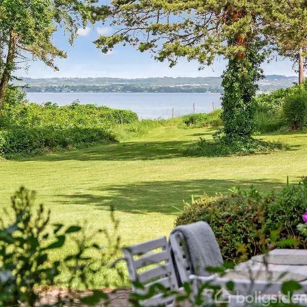 En have med en hvid havebordsgruppe står foran et græsklædt område, der strækker sig mod en sø omgivet af træer og buskads. I baggrunden ses en statue.