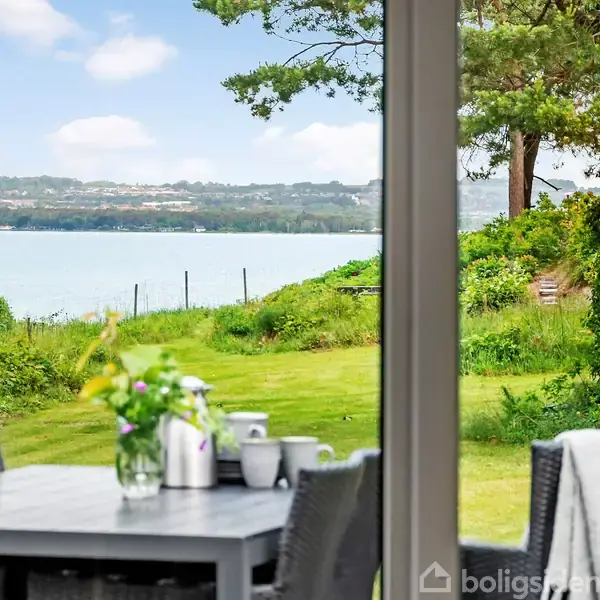 Bord med blomsterarrangement og kaffekopper står ude i en have. Udsigt over græsplæne mod en sø, omkranset af grønne buske og træer.