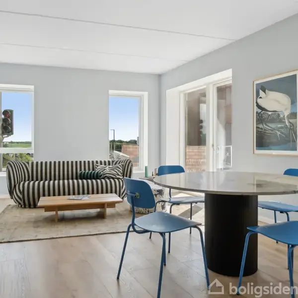 En stribet sofa står ved et sofabord i en moderne stue med lysegrå vægge. Fire blå stole omgiver et rundt spisebord. Store vinduer giver udsigt til naturen udenfor.