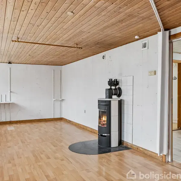 Et rum med trægulv og træloft har en fritstående brændeovn mod en hvid væg. Rummet er lyst med flere vinduer og har en åbning til et tilstødende køkken med hvide elementer.