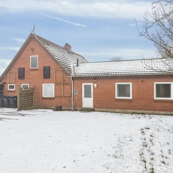 Rødt murstenshus står stille i et sneklædt landskab med en blå himmel. Huset har flere vinduer og en hvid dør. Træer og et hegn omgiver bygningen.