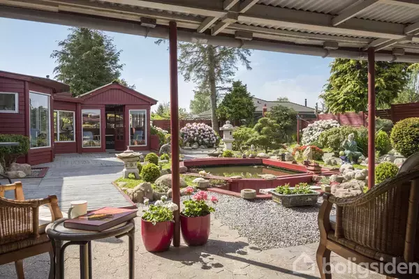 En overdækket terrasse med stole og borde, vender ud mod en have med sten, blomster og et lille vandbassin. Omkring haven ses røde bygninger og grønne træer samt buske.