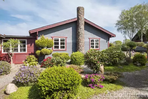 Et hyggeligt hus med blå og rød facade har en central stenskorsten. Foran ses en velplejet have med buske og blomster, omgivet af en grussti, under en blå himmel.