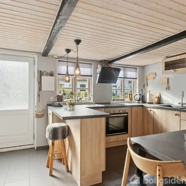 Køkkenbord med indbygget kogeplade og emhætte, omgivet af træskabe og stole, i et lyst rum med store vinduer, loftsbjælker og dekorative lamper, ved siden af en glasdør mod venstre.