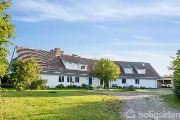 En hvid bygning med rødt tegltag står stille i en grøn have. Gruset indkørsel fører op til huset, som er omgivet af træer og buske under en klar blå himmel.