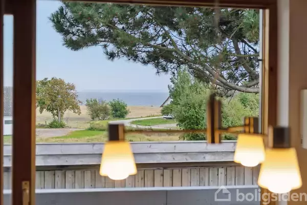 Lamper hænger fra loftet, oplyser et rum med havudsigt. Udenfor er der træer, en grusvej og havet i horisonten, set gennem vinduet.