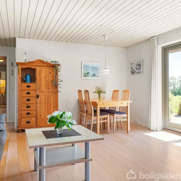 En lys stue med trægulv indeholder en grå sofa og et sofabord med blomster. Et spisebord med stole står foran en stor glasdør, der viser en grøn have udenfor.