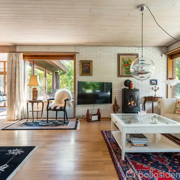 Stue med moderne indretning; pejs brænder ved siden af hvide sofabord og sofa med puder. Store vinduer lader naturligt lys strømme ind, omgivet af grønne omgivelser. Malerier hænger på væggene.