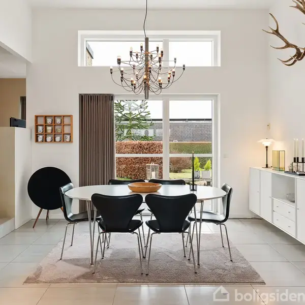 Spisebord med seks stole står på et tæppe i en lys spisestue. En moderne lysekrone hænger over bordet. Rummet har loftvindue, dekorative horn og grønne planter.