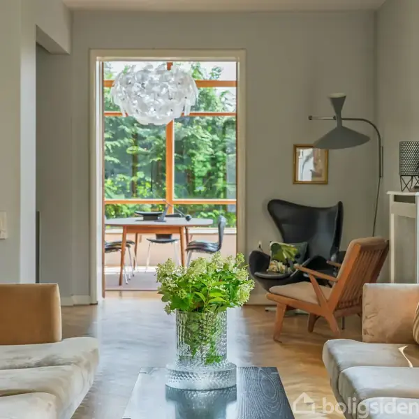 Murstenshus med høje vinduer og en trappe fører ned til en terrasse. I en have står et hvidt bord og stol på flisebelægning omgivet af græs og buske.