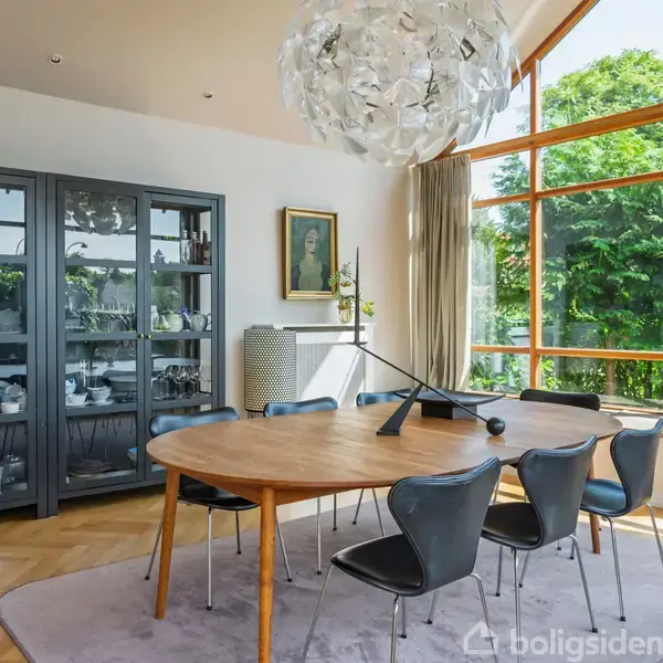 Rundt spisebord i træ står i en lys spisestue med store vinduer. Seks stole omgiver bordet. Et stort, kunstnerisk loftlys hænger over bordet. Skab med servise i baggrunden.