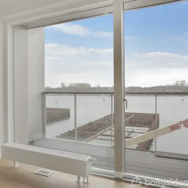 Glasdør åbner ud til en balkon med udsigt over en rolig vandflade og skyet himmel. Indeni ses lyst trægulv og en radiator.