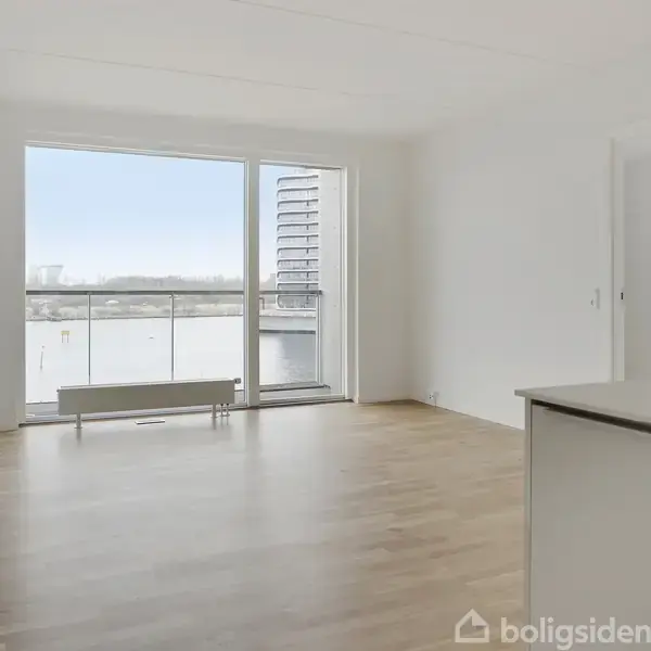 Et lyst rum med trægulv, store vinduer og en altandør. Udsigt til en balkon og en moderne bygning udenfor. Enkel, hvid væg og begrænset møblering.