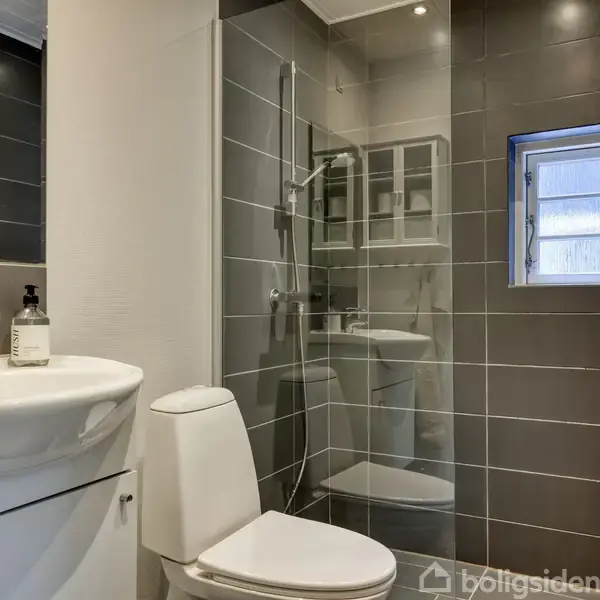 Hvidt badeværelse med toilet, håndvask og brusekabine. Vægge er delvist flisebelagte i gråt, og der er et lille vindue ved bruseren. Der står en sæbeflaske ved håndvasken.
