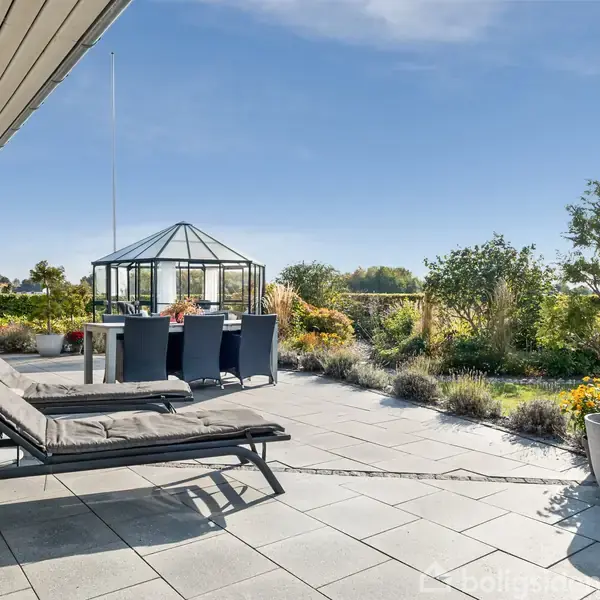 To liggestole står på en terrasse med flisebelægning. I baggrunden ses et spisebord med stole samt en have omkranset af buske og blomster under en blå himmel.