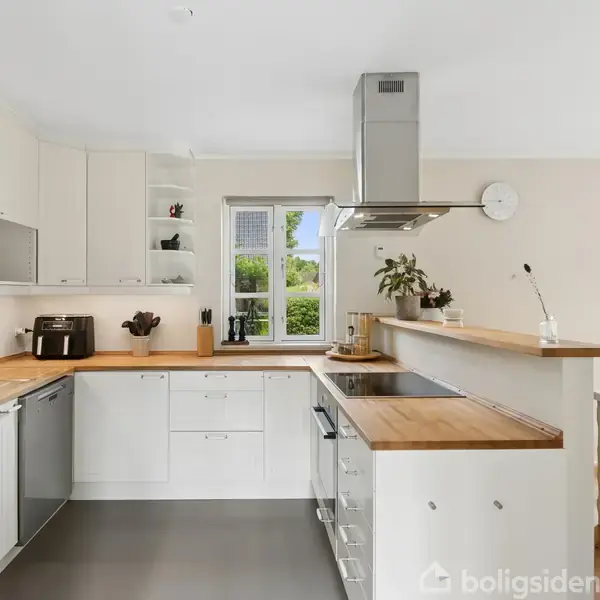 Køkken med hvide skabe og træbordplader, udstyret med moderne apparater som køleskab og ovn. En plante står på bordet, og en spiseplads kan ses i baggrunden ved et stort vindue.
