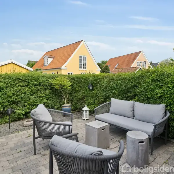 Havemøbler i grå betræk står på en flisebelagt terrasse. Handlingen er ro og afslapning med en grøn hæk omkring. Gule huse ses i baggrunden under en blå himmel.