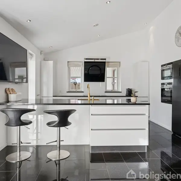 Et moderne køkken med en stor, sort flisebelagt ø. To høje sorte stole står ved øen. Omkring ses et sort køleskab, ovn og en vægmonteret fladskærm.