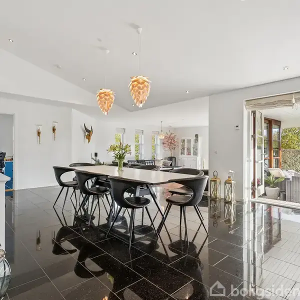 En moderne spisestue med et sort bord og seks sorte stole. Rummet har sort glansgulv og tre hængende lampeskærme. Store åbne døre fører ud til en terrasse med grønne omgivelser.