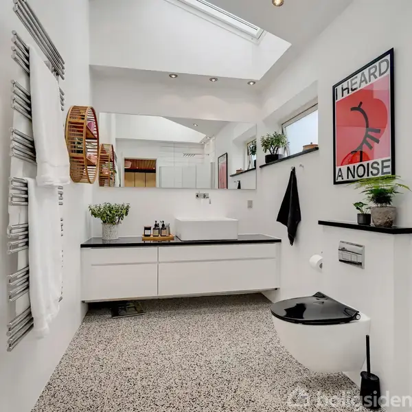 Badeværelse med hvidt interiør, en håndvask på stor bordplade, vægmonteret toilet og tekstplakat med "HEARD A NOISE". Vægge og gulv har en lys farve, dekorativ radiator til håndklæder.