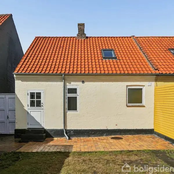 Hus med hvid facade og orange tegltag ses bagfra. Dør og vindue på bagsiden. Gårdsplads med fliser og lille græsplæne omringet af gult plankeværk.