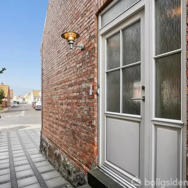 Dør med glasruder står lukket på en smal fortov med murstensvægge på begge sider i en bymæssig gade. En udendørs lampe er monteret over døren.