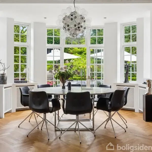 En elegant trappe fører opad i en lys gang med parketgulv. En sort lænestol står ved siden af et TV, omgivet af runde gyldne spejle og en moderne lampe.