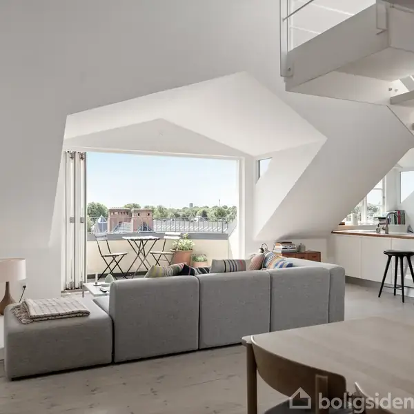 Moderne stue med grå sofa, hvor en person sidder. Store vinduer giver udsigt til byen. Til højre er der et åbent køkken og en trætrappe op til en etage.