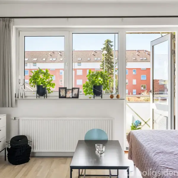 Et værelse møbleret med en kommode, fladskærms-TV og en seng; et bord står foran vinduer, hvor planter og billeder er placeret, udsigt til røde bygninger udenfor.