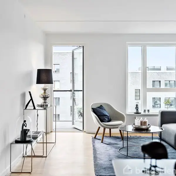 Stuen har en grå sofa og lænestol ved et vindue, med et rundt sofabord. Der er lyse vægge, to arkitektlamper, en reol med dekorationer og udsigt til bygninger udenfor.