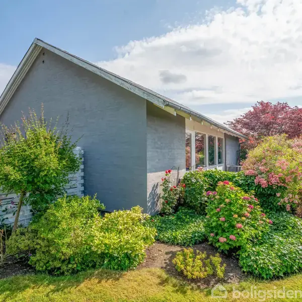 Et gråt hus med gavltag omgivet af frodig have med forskellige blomstrende buske og træer. Huset har en række vinduer mod haven under en skyfri blå himmel.