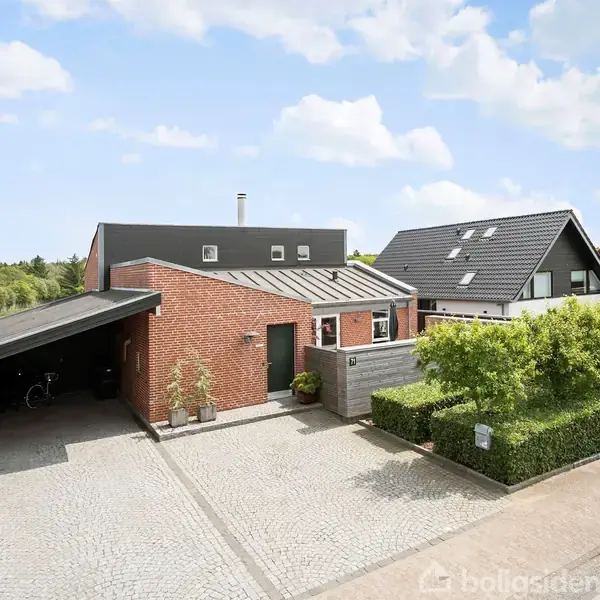 Et moderne, rødt murstenshus med fladt tag står foran en brolagt indkørsel, ved siden af en carport med cykler. Omgivet af grønne buske og nabohuse i et boligområde.