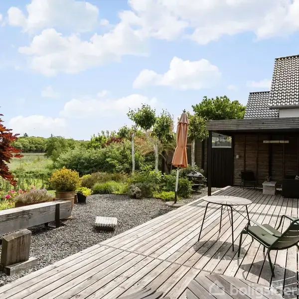 Træterrasse med to stole og et bord, omgivet af blomster og buske. En orange parasol står ved siden af terrassen. I baggrunden ses en græsplæne og blå himmel med skyer.