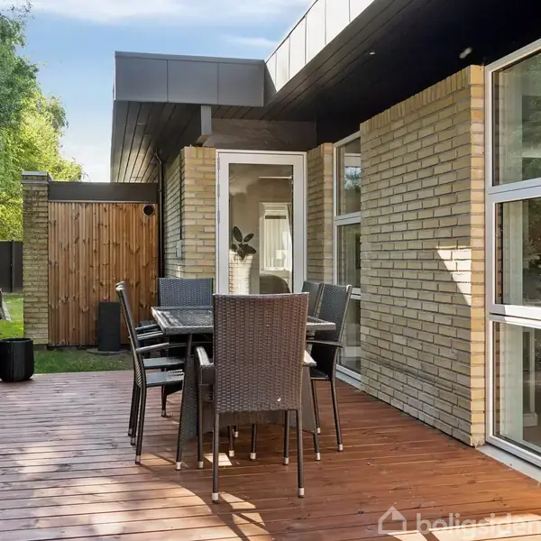 En terrasse med et bord og seks stole af mørkt materiale. Terrassen er lavet af træplanker og ligger op ad et murstenshus med store vinduer. Omkring er der en velplejet hæk og træer.