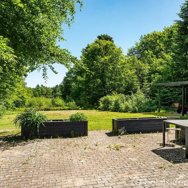 Terrasse med flisebelægning omgivet af grønne planter. Lige foran står et havebord med stole. I baggrunden ses en frodig have og træer under en klar blå himmel.