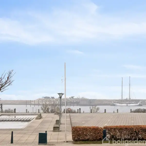 En åben plads med fliser omgivet af lave busker og et træ; i baggrunden ses en havnefront med fortøjede sejlbåde under en blå himmel.