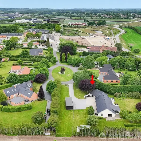 En gruppe af huse er omgivet af grønne haver. En rød pil peger på et hus i midten. Landskabet er opdelt med veje og marker i baggrunden.