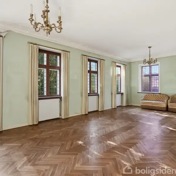 En stor, tom stue med parketgulv, lysekroner og flere store vinduer med gardiner. To beige sofaer står i hjørnet, og der er grønne vægge. Ingen tekst er synlig.
