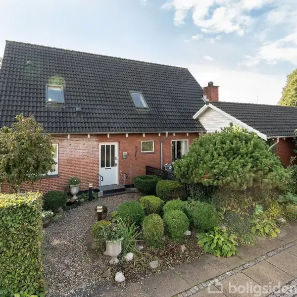 Et rødt murstenshus med sort tag står omkranset af velplejede buske og træer. En smal grussti fører til hoveddøren. Huset har to etager med skrå vinduer.