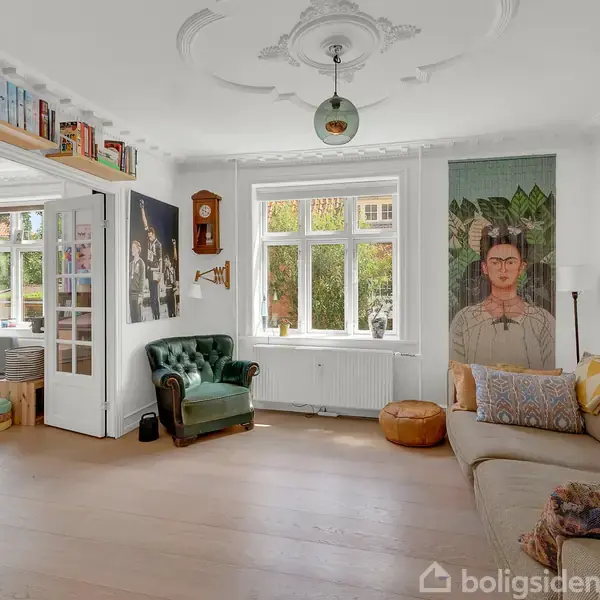 Stue med sofa til højre, grøn lænestol til venstre, og vægdekorationer. Store vinduer lader lys ind. Bogreol på væggen og en dør åbner til et tilstødende rum.