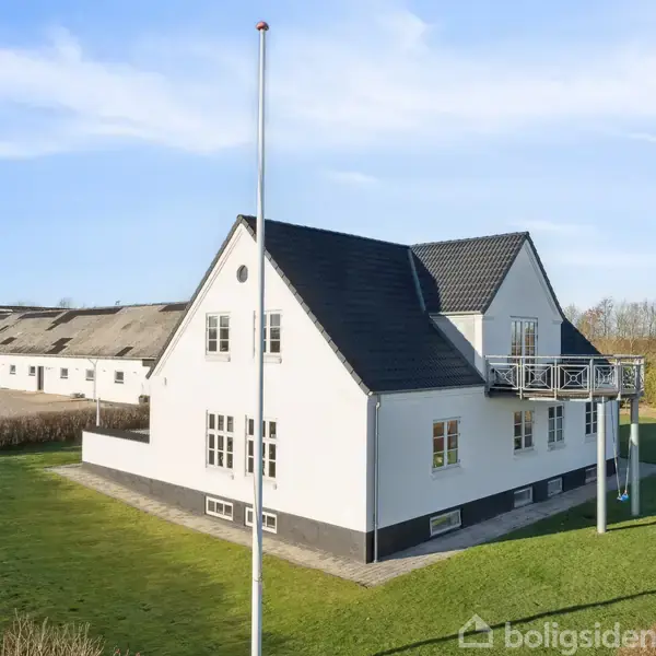 Et hvidt to-etagers hus med sort tag står på en velplejet græsplæne. En flagstang står foran huset, der er omgivet af buske, med en lade i baggrunden.