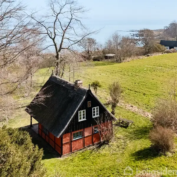 Et bindingsværkshus med stråtag står på en grøn mark nær kysten. Træer omkranser området, og der er udsigt til havet og nogle spredte huse i baggrunden.