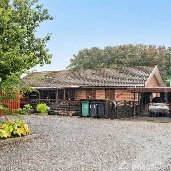 En etplans stenhus med fladt tag, omgivet af grusbelagt gårdsplads og grønne planter. En bil er parkeret under en carport ved huset.