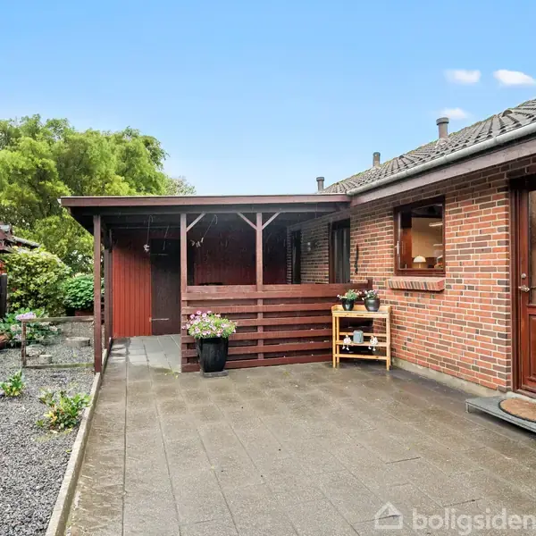 En rød murstensbygning med en trædør åbner ud til en flisebelagt terrasse. Der står planter i krukker. Til venstre er der et stakit, og i baggrunden ses grønne træer.
