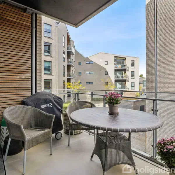En balkon med et rundt bord i midten, omgivet af to kurvestole og blomster. En grill står i hjørnet. Udsigt til moderne lejlighedsbygninger.
