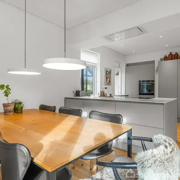 Et træbord med sorte stole står i et moderne køkken-alrum. Bordet er pyntet med en plante. Bagved ses et køkken med grå skabe og køkkenø, oplyst af loftslamper.