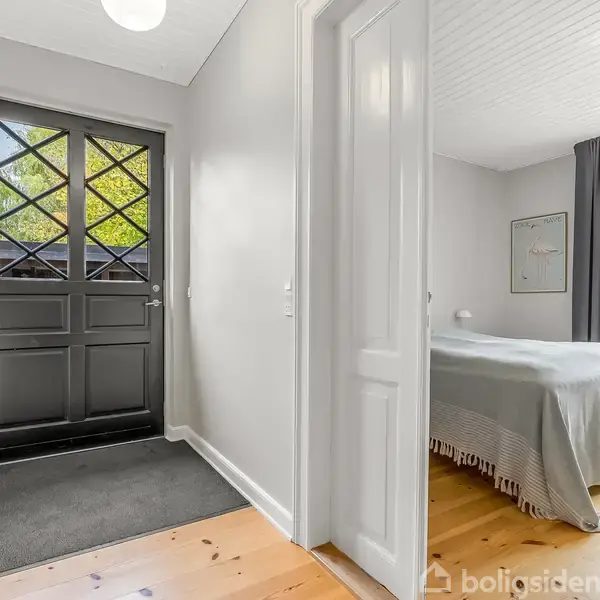 Sort dør med glasruder åbner ind til en lys gang med trægulv. Til højre ses en åben dør til et soveværelse med dobbelt seng og grå gardiner.