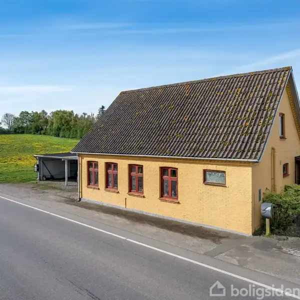 Gult murstenshus står stille ved landevej, omgivet af marker og en gårdsplads. Taget er mørkt og skrånende. En række rødrammede vinduer vender mod vejen.