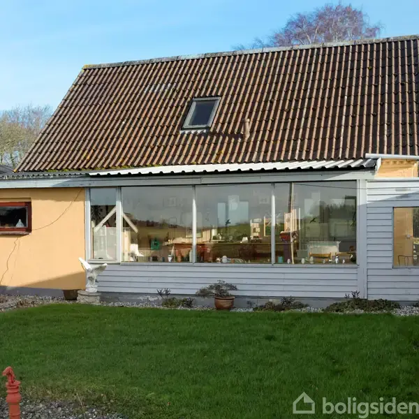 Et hus med skråtag er omgivet af en græsplæne og fliser. Store vinduer åbner mod haven, og en rød bygning står ved siden af. Spredte planter ses foran huset.