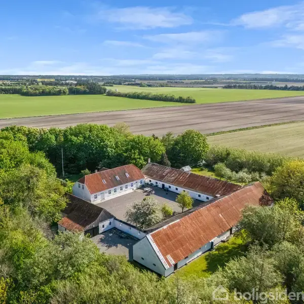 Gårdkompleks med røde tage ligger omgivet af grønne marker og tæt vegetation, danner en firkantet gårdsplads. I baggrunden strækker landbrugslandskabet sig under en blå himmel.